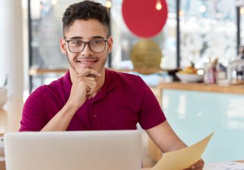 Satisfied man works on clients project, updates software, sits in front of opened laptop computer, holds document, keeps hand on chin, models over cozy cafeteria interior, thinks on idea for startup